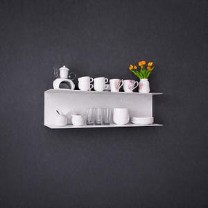 K&ouml;nnte beinhalten: Ein wei&szlig;es, an der Wand befestigtes Regal zeigt eine Sammlung wei&szlig;er Keramikgeschirr. Dazu geh&ouml;ren Teetassen, eine Teekanne, Schalen und Gl&auml;ser. Eine kleine Vase mit orangefarbenen Blumen sorgt f&uuml;r einen Farbtupfer. Das Regal befindet sich vor einer dunkelgrauen Wand.