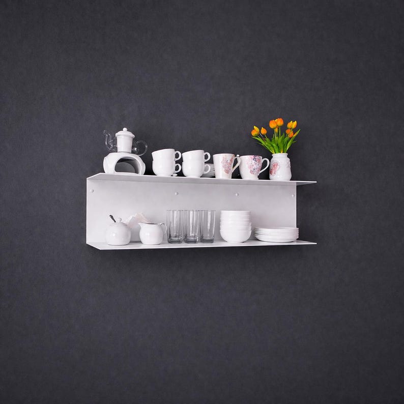 K&ouml;nnte beinhalten: Ein wei&szlig;es, an der Wand befestigtes Regal zeigt eine Sammlung wei&szlig;er Keramikgeschirr. Dazu geh&ouml;ren Teetassen, eine Teekanne, Schalen und Gl&auml;ser. Eine kleine Vase mit orangefarbenen Blumen sorgt f&uuml;r einen Farbtupfer. Das Regal befindet sich vor einer dunkelgrauen Wand.