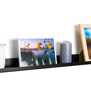 May include: A black metal shelf with a curved edge, mounted on a white wall. The shelf is displaying a white picture frame with a photo of yellow flowers, a gray candle, and a wooden picture frame with a photo of a tall grass.