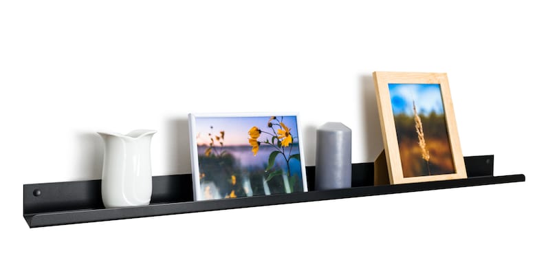 May include: A black metal shelf with a curved edge, mounted on a white wall. The shelf is displaying a white picture frame with a photo of yellow flowers, a gray candle, and a wooden picture frame with a photo of a tall grass.
