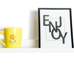 May include: A white shelf with a yellow mug with a flower design, a black picture frame with the word "ENJOY" in white letters, and a gray candle.