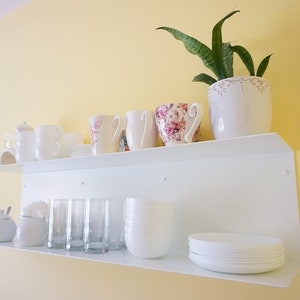 K&ouml;nnte beinhalten: Ein wei&szlig;es Wandregal zeigt verschiedene K&uuml;chenartikel. Das obere Regal enth&auml;lt wei&szlig;e Tassen, eine Teekanne und eine Tasse mit Blumenmuster. Das untere Regal bietet Gl&auml;ser, Schalen und Teller, mit einer kleinen Pflanze in einem wei&szlig;en Topf.