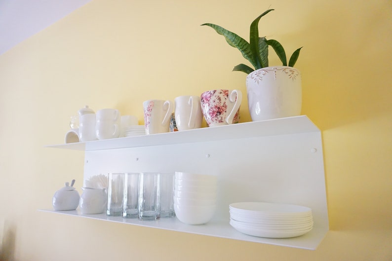 K&ouml;nnte beinhalten: Ein wei&szlig;es Wandregal zeigt verschiedene K&uuml;chenartikel. Das obere Regal enth&auml;lt wei&szlig;e Tassen, eine Teekanne und eine Tasse mit Blumenmuster. Das untere Regal bietet Gl&auml;ser, Schalen und Teller, mit einer kleinen Pflanze in einem wei&szlig;en Topf.