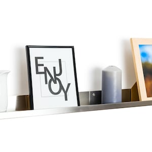 May include: A silver metal shelf with a white ceramic pitcher, a framed print with the word "ENJOY" in black lettering, a gray candle, and a framed photograph of a tall grass stalk against a blue sky.
