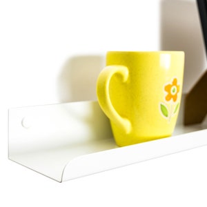 May include: A white metal shelf with a rounded edge, mounted to a white wall. A yellow mug with a flower design sits on the shelf.