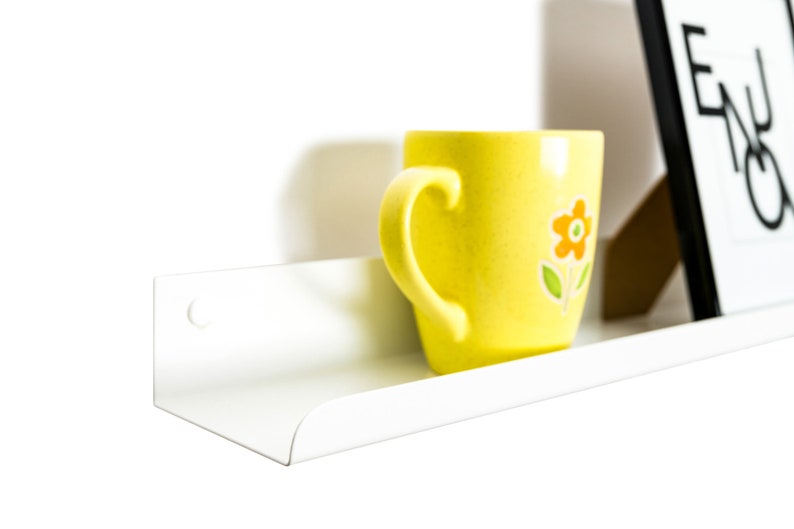 May include: A white metal shelf with a rounded edge, mounted to a white wall. A yellow mug with a flower design sits on the shelf.