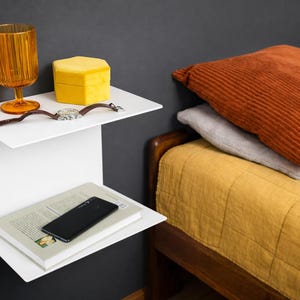 K&ouml;nnte beinhalten: Ein wei&szlig;es, an der Wand befestigtes Regal mit zwei Ebenen. Auf der oberen Ablage befinden sich ein orangefarbenes Glas, eine gelbe Schachtel und eine Armbanduhr. Auf der unteren Ablage liegen ein Buch und ein schwarzes Smartphone. Im Hintergrund ein Bett mit orangefarbenen und grauen Kissen.