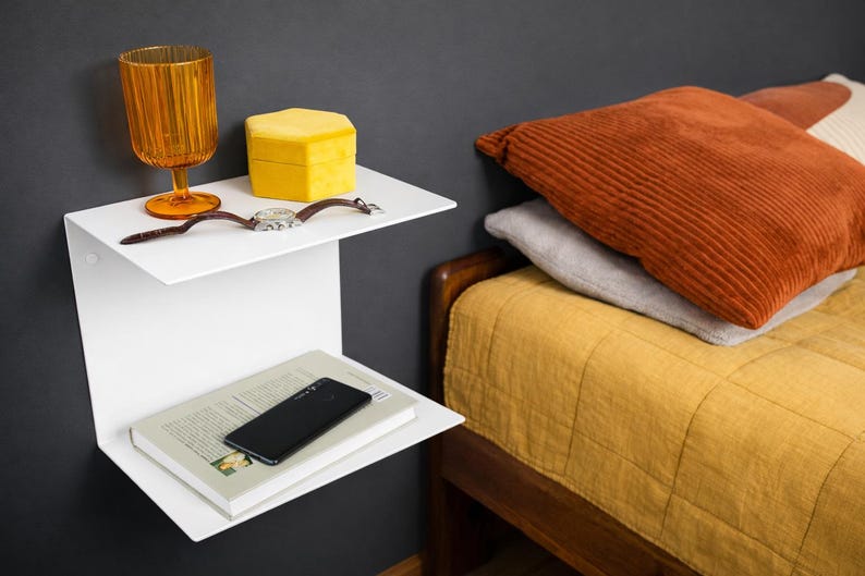 K&ouml;nnte beinhalten: Ein wei&szlig;es, an der Wand befestigtes Regal mit zwei Ebenen. Auf der oberen Ablage befinden sich ein orangefarbenes Glas, eine gelbe Schachtel und eine Armbanduhr. Auf der unteren Ablage liegen ein Buch und ein schwarzes Smartphone. Im Hintergrund ein Bett mit orangefarbenen und grauen Kissen.