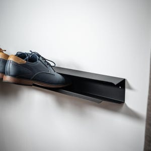 Peut inclure: Un porte-chaussures en m&eacute;tal noir mont&eacute; sur un mur blanc avec une paire de chaussures en cuir bleu sur le porte-chaussures.