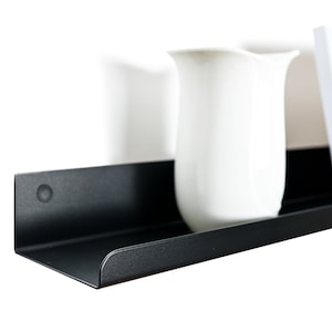 May include: A black metal shelf with rounded edges, featuring a white pitcher and a framed photograph with a landscape scene.