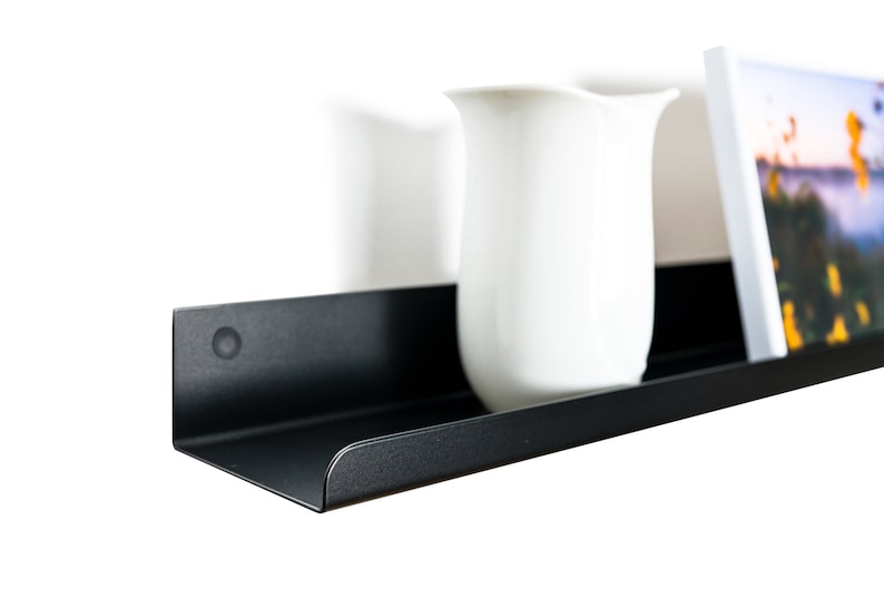 May include: A black metal shelf with rounded edges, featuring a white pitcher and a framed photograph with a landscape scene.
