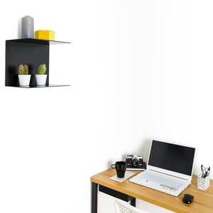 K&ouml;nnte beinhalten: Ein modernes Homeoffice-Setup mit einem hellen Holzschreibtisch und einem schwarzen Metallrahmen. Ein wei&szlig;er Laptop, eine schwarze Kaffeetasse und eine Vintage-Kamera stehen auf dem Schreibtisch. Ein schwarzes Wandregal zeigt Kakteen, eine gelbe Box und eine graue Kerze.