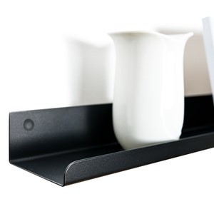 May include: A black metal shelf with rounded edges, featuring a white pitcher and a framed photograph with a landscape scene.