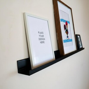 Op de afbeelding: Een zwarte metalen zwevende plank toont ingelijste kunstwerken. Een wit ingelijste print leest "PLACE YOUR DESIGN HERE". Een bruin ingelijste print toont een hanenontwerp. Een kleine zwart ingelijste print.