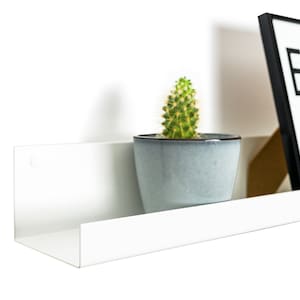 Modern floating metal picture ledge made of 1.5 mm cold-rolled steel, with rounded corners and front lip. Ideal for framed photos, art prints, books, spices or decor. Powder-coated in black, black matt, white, white matt, or brushed stainless steel for industrial, minimalist, or Scandinavian interiors. Wall-mounted with hidden hardware. Available in custom RAL colors and lengths up to 240 cm.