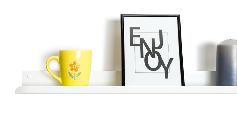 May include: A white shelf with a yellow mug with a flower design, a black picture frame with the word "ENJOY" in white letters, and a gray candle.