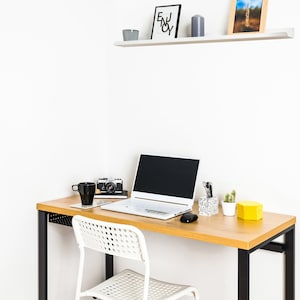 May include: A white laptop computer with a black keyboard and a black screen is on a light wood desk with a black metal frame. A white chair with a black metal frame is next to the desk. There is a black camera, a white mug, and a gray candle on the desk. There is a white shelf above the desk with a black framed print that says "Enjoy" and a brown framed picture.