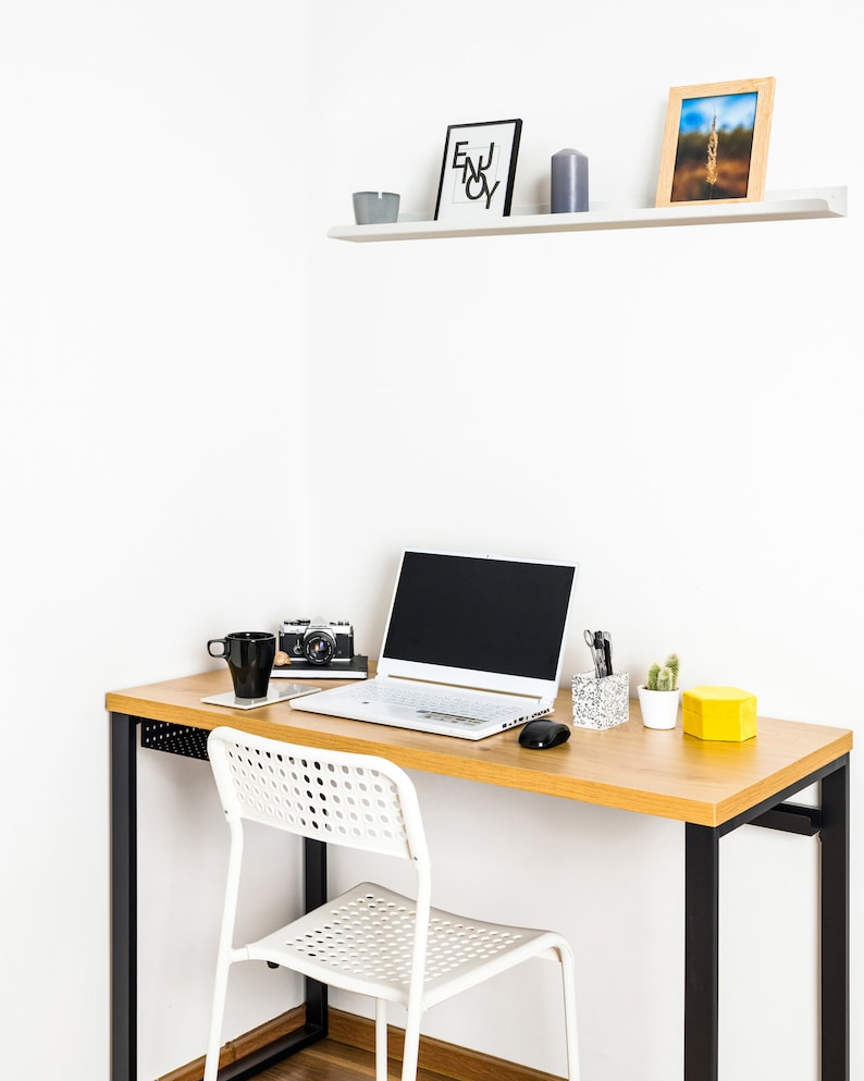 May include: A white laptop computer with a black keyboard and a black screen is on a light wood desk with a black metal frame. A white chair with a black metal frame is next to the desk. There is a black camera, a white mug, and a gray candle on the desk. There is a white shelf above the desk with a black framed print that says "Enjoy" and a brown framed picture.