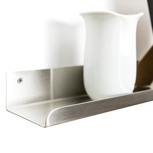 May include: A silver metal shelf with a rounded edge, mounted to a white wall. A white pitcher sits on the shelf.
