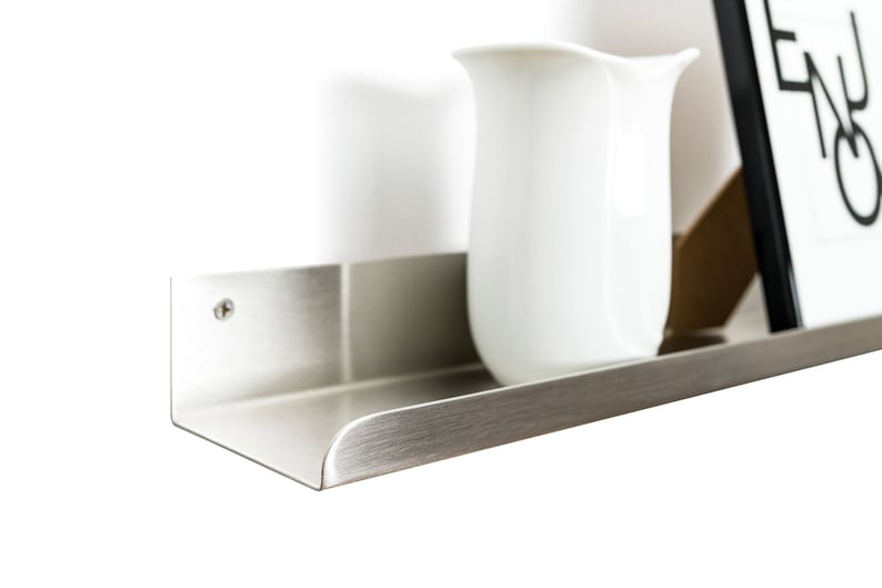 May include: A silver metal shelf with a rounded edge, mounted to a white wall. A white pitcher sits on the shelf.