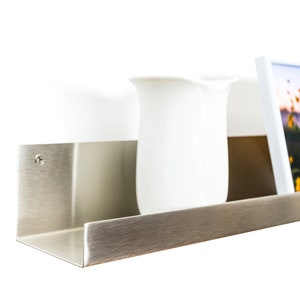 Modern floating metal picture ledge made of 1.5 mm cold-rolled steel, with rounded corners and front lip. Ideal for framed photos, art prints, books, spices or decor. Powder-coated in black, black matt, white, white matt, or brushed stainless steel for industrial, minimalist, or Scandinavian interiors. Wall-mounted with hidden hardware. Available in custom RAL colors and lengths up to 240 cm.