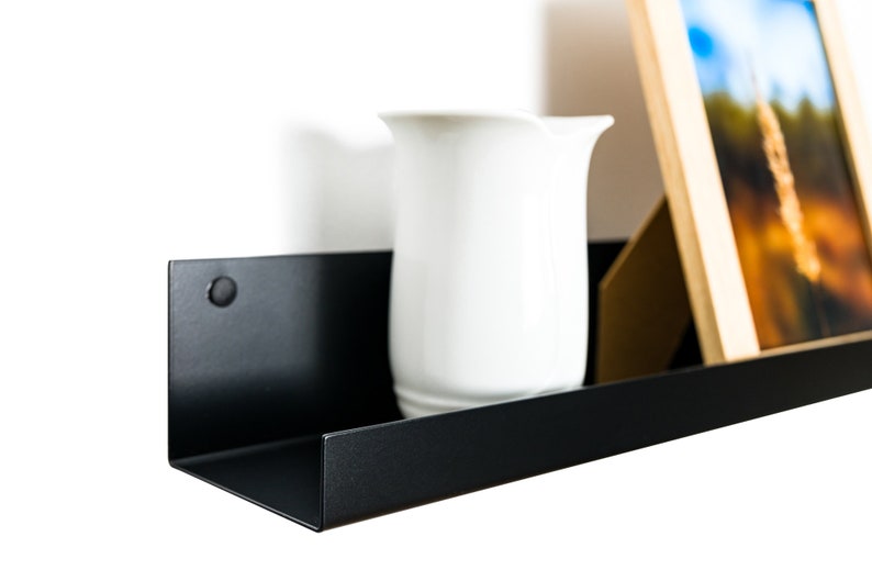 Modern floating metal picture ledge made of 1.5 mm cold-rolled steel, with rounded corners and front lip. Ideal for framed photos, art prints, books, spices or decor. Powder-coated in black, black matt, white, white matt, or brushed stainless steel for industrial, minimalist, or Scandinavian interiors. Wall-mounted with hidden hardware. Available in custom RAL colors and lengths up to 240 cm.
