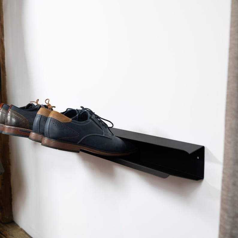 Peut inclure: Un porte-chaussures en m&eacute;tal noir mont&eacute; sur un mur blanc avec deux paires de chaussures dessus. Les chaussures sont une paire en cuir bleu fonc&eacute; et une paire en cuir marron.
