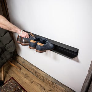 Peut inclure: Un porte-chaussures en m&eacute;tal noir mont&eacute; sur un mur blanc. Le porte-chaussures a un bord incurv&eacute; et est con&ccedil;u pour tenir les chaussures horizontalement. Trois paires de chaussures sont affich&eacute;es sur le porte-chaussures, dont une paire de chaussures en cuir marron et deux paires de chaussures en cuir bleu.