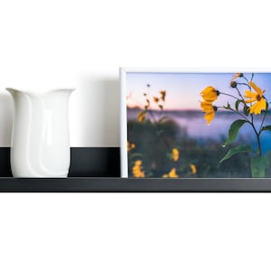 May include: A black metal shelf with a white vase, a framed photograph of yellow flowers, and a gray candle. The shelf is mounted on a white wall.