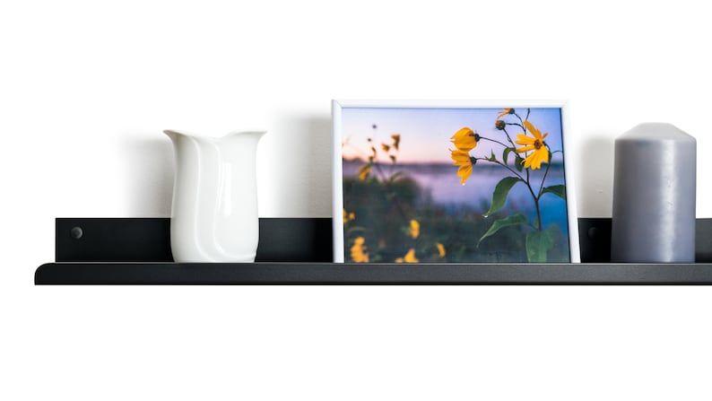 May include: A black metal shelf with a white vase, a framed photograph of yellow flowers, and a gray candle. The shelf is mounted on a white wall.