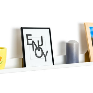 May include: White floating shelf with a yellow mug with a flower design, a black framed print with the word "ENJOY" in white, a gray candle, and a wooden framed photo with a landscape scene.