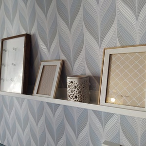 May include: Four picture frames and a white ceramic candle holder on a white shelf against a blue and white patterned wallpaper. The picture frames are all different sizes and styles. The candle holder has a decorative design. The largest picture frame has the text "13x18cm" printed on the bottom.