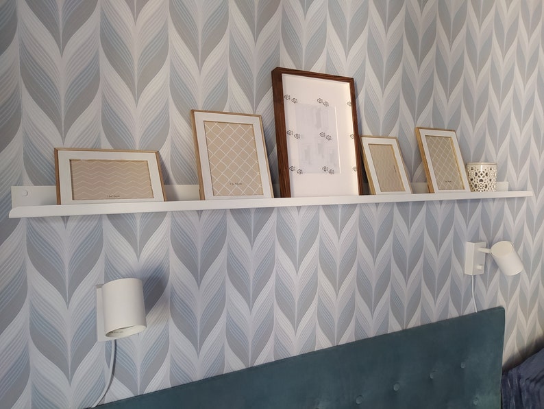 May include: A white shelf with five framed pictures and a small candle holder. The shelf is mounted on a wall with a light blue and gray patterned wallpaper. There are two white wall sconces on the wall.