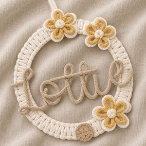 May include: A cream and tan macrame wreath with the word "little" in cursive. The wreath is decorated with three yellow and cream crocheted flowers. A small wooden tag at the bottom reads "Handmade by Pretty Paper Play."