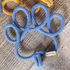 May include: A blue knitted coaster in a clover shape, with the text "Wire & Whimsy" on a small tag. The coaster is made of thick, textured yarn. A yellow knitted item is partially visible in the background.