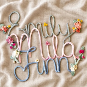 May include: A wall decoration featuring the names Emily, Mila, and Finn written in cursive script using knitted rope. The names are in shades of grey, pink, and blue, accented with small, colourful crocheted flowers and green leaves.