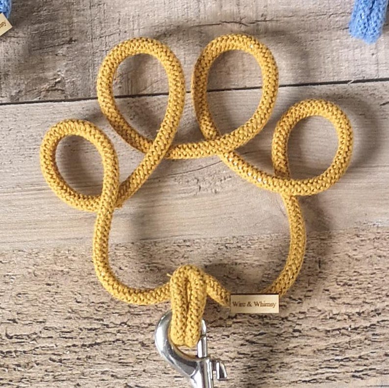 May include: Mustard yellow dog leash in the shape of a paw print. The leash is made of a thick, woven material and has a silver metal clasp. A small wooden tag with the text "Ware & Whimsy" is attached.
