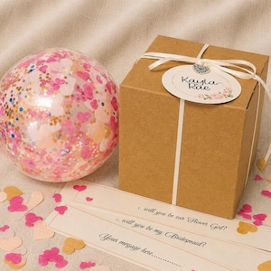 May include: A clear balloon filled with pink and gold heart-shaped confetti next to a square brown cardboard gift box tied with a white ribbon. A round tag attached to the ribbon reads Kayla-Rae. Three paper strips feature the text: will you be our Flower Girl?, will you be my Bridesmaid?, and Your message here.