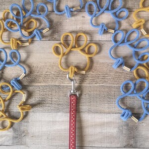 May include: A dog leash with a red and white polka dot pattern is attached to a gold paw-shaped rope. Surrounding the leash are blue and gold rope paw prints, each with a small tag. The items are arranged on a wooden surface.