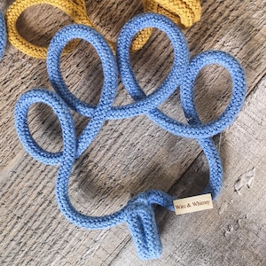 May include: A close-up of a blue knitted coaster in a clover shape. The coaster is made of thick, soft yarn and has a tag that reads "Wire & Whimsy". A yellow knitted coaster is partially visible in the background.