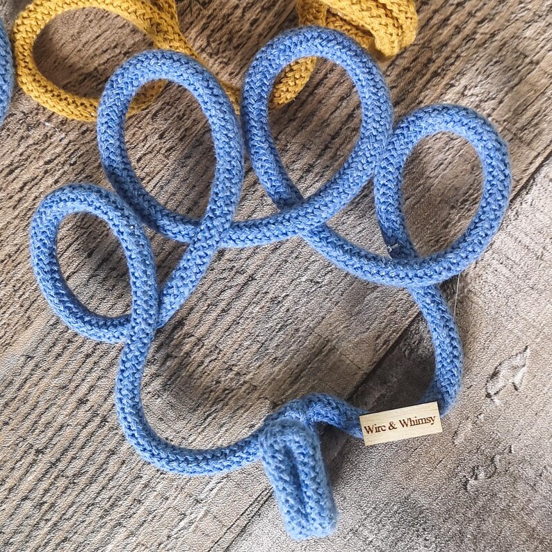 May include: A close-up of a blue knitted coaster in a clover shape. The coaster is made of thick, soft yarn and has a tag that reads "Wire & Whimsy". A yellow knitted coaster is partially visible in the background.