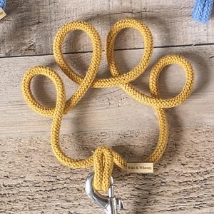 May include: Mustard yellow dog leash in the shape of a paw print. The leash is made of a thick, woven material and has a silver-colored metal clasp. The tag reads "Ware & Whimsy".