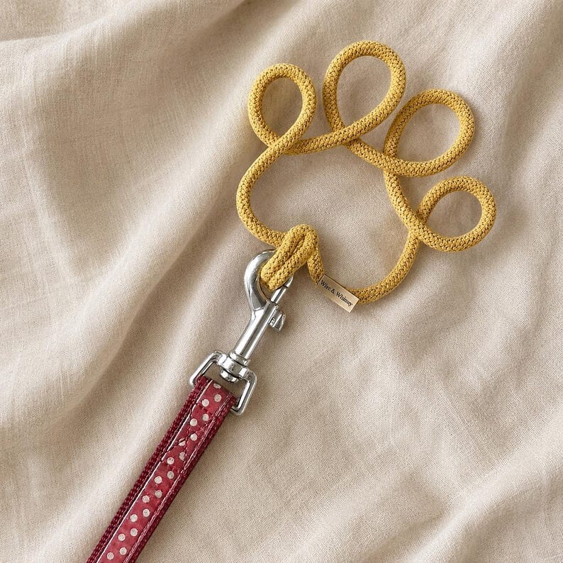May include: A dog leash with a red and white polka dot pattern. The leash connects to a yellow rope paw print design with a silver metal clasp. The paw print has a small tag that reads "Wife & Whimsy".