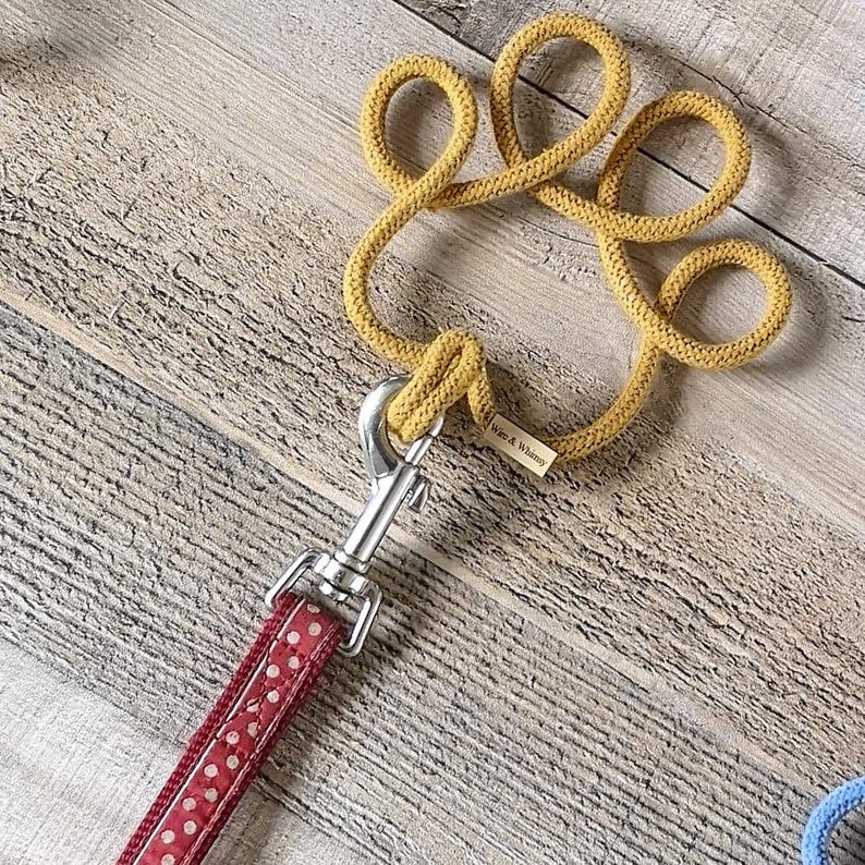 May include: A mustard yellow rope dog leash shaped like a paw, connected to a red polka dot leash. The leash has a silver metal clasp. The tag reads "Worn & Whimsy". The background is a light brown wood.