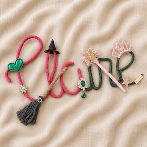 May include: A decorative name sign spelled out in cursive rope, featuring a pink rope section with a green heart, a small black witch hat, and a broom, and a dark green rope section with a pink star wand, a pink crown, and a green jewel brooch.