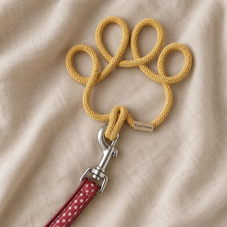 May include: A yellow rope dog leash shaped like a paw print. The leash has a silver metal clasp and a red and white polka dot patterned handle. The tag reads "Wag & Whimsy".