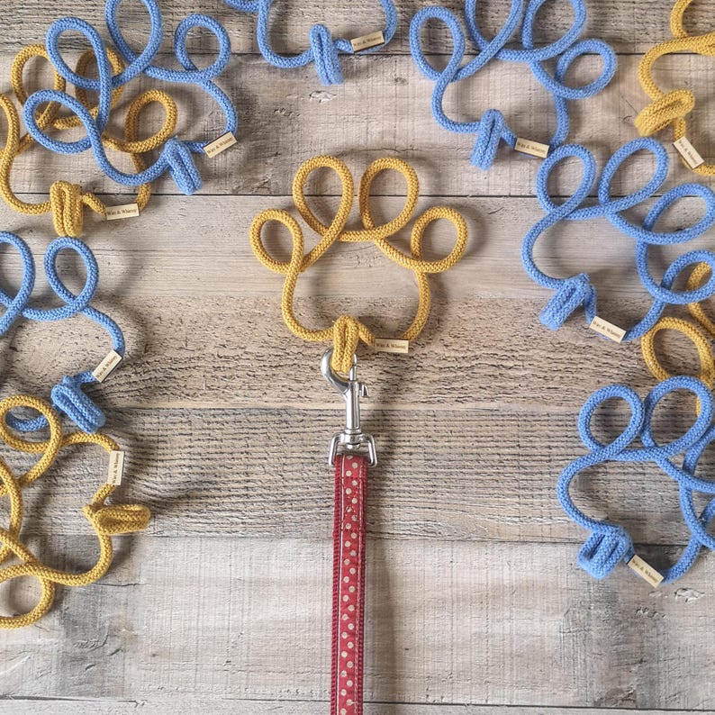May include: A dog leash with a red and white polka dot pattern is attached to a gold paw-shaped rope. Surrounding the leash are blue and gold rope paw prints, each with a small tag. The items are arranged on a wooden surface.