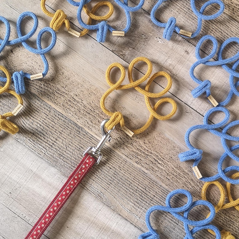 May include: A red polka-dot dog leash with a silver clasp is attached to a yellow rope dog toy. Several blue and yellow rope toys are scattered on a wooden surface. Each toy has a small wooden tag.