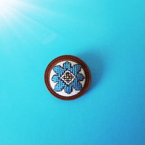 Könnte beinhalten: Ein runder Holz-Ansteckknopf mit einem gestickten Blumenmuster in Blau und Weiß.