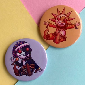 May include: Two round pinback buttons featuring cartoon jester characters. The button on the left has a purple background with a black jester with red eyes and a white star on its hat. The button on the right has an orange background with a red jester with yellow eyes and a yellow star on its hat.